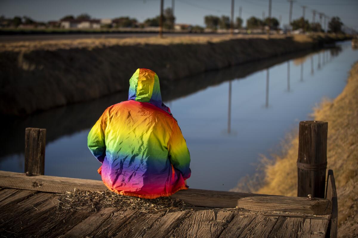A young man in a rainbow colored jacket sits on a wooden dock near a canal