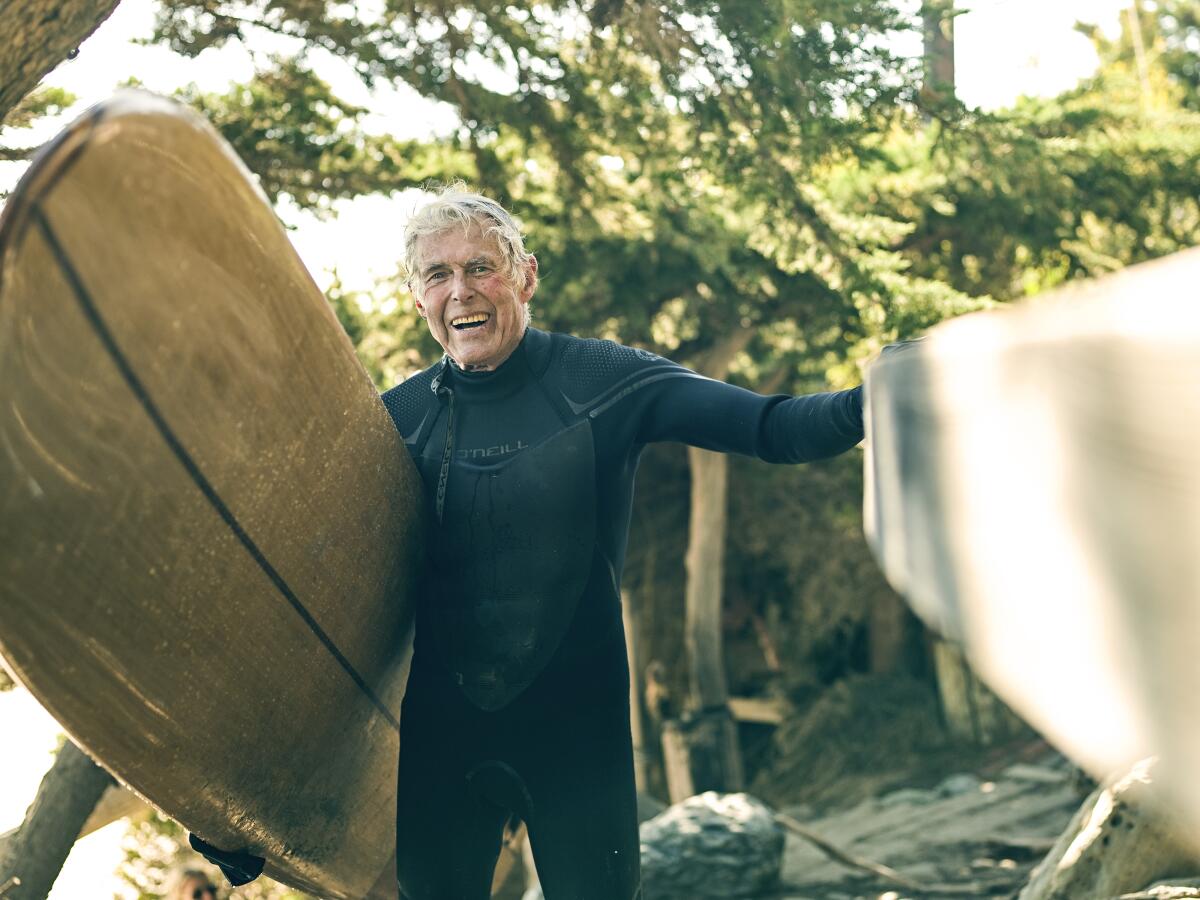 Harry Gesner is seen in a black wetsuit holding a surfboard