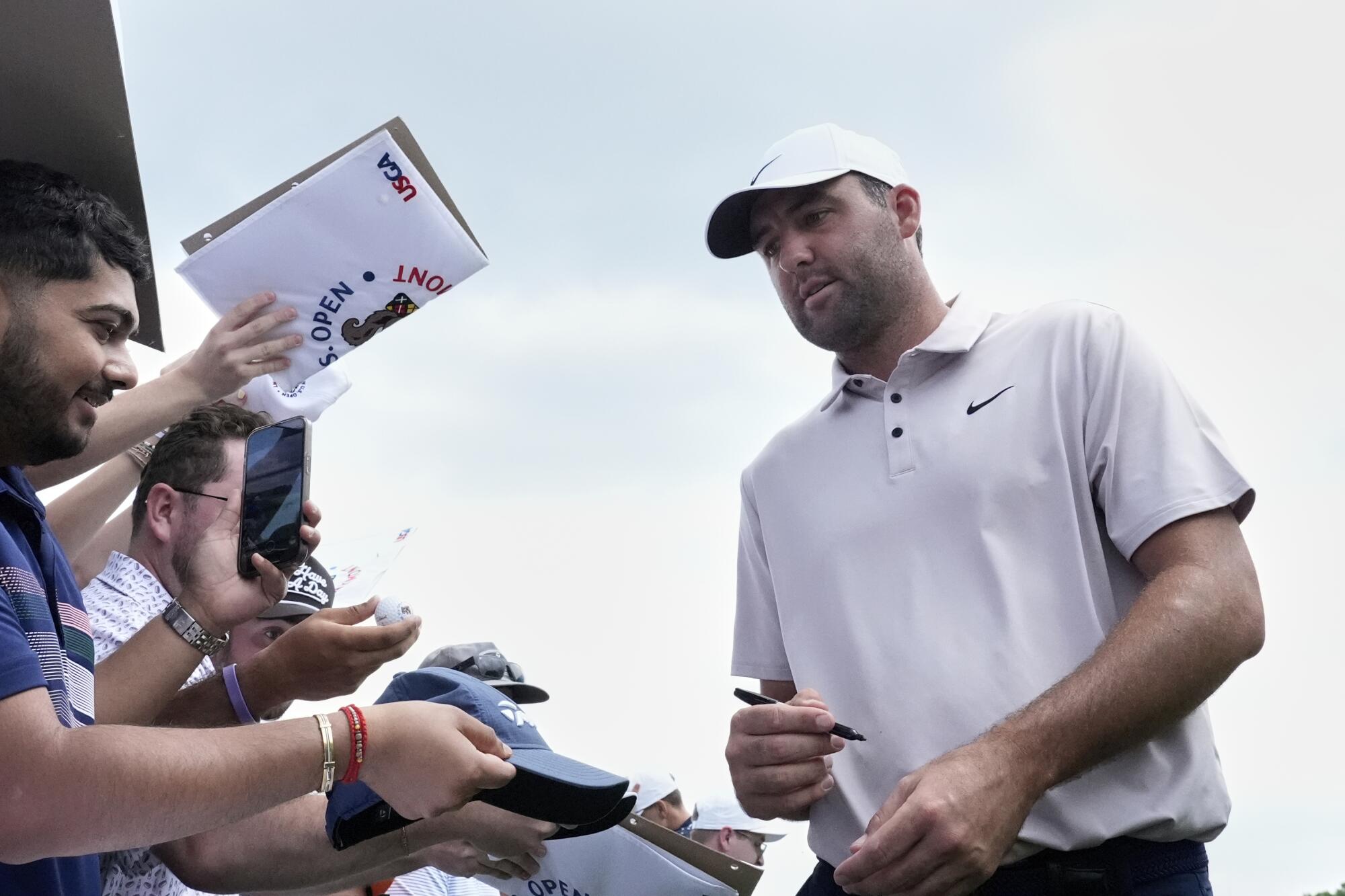 Scottie Scheffler in a white golf shirt stands in front of a group of fans with a pen in his hand to sign autographs