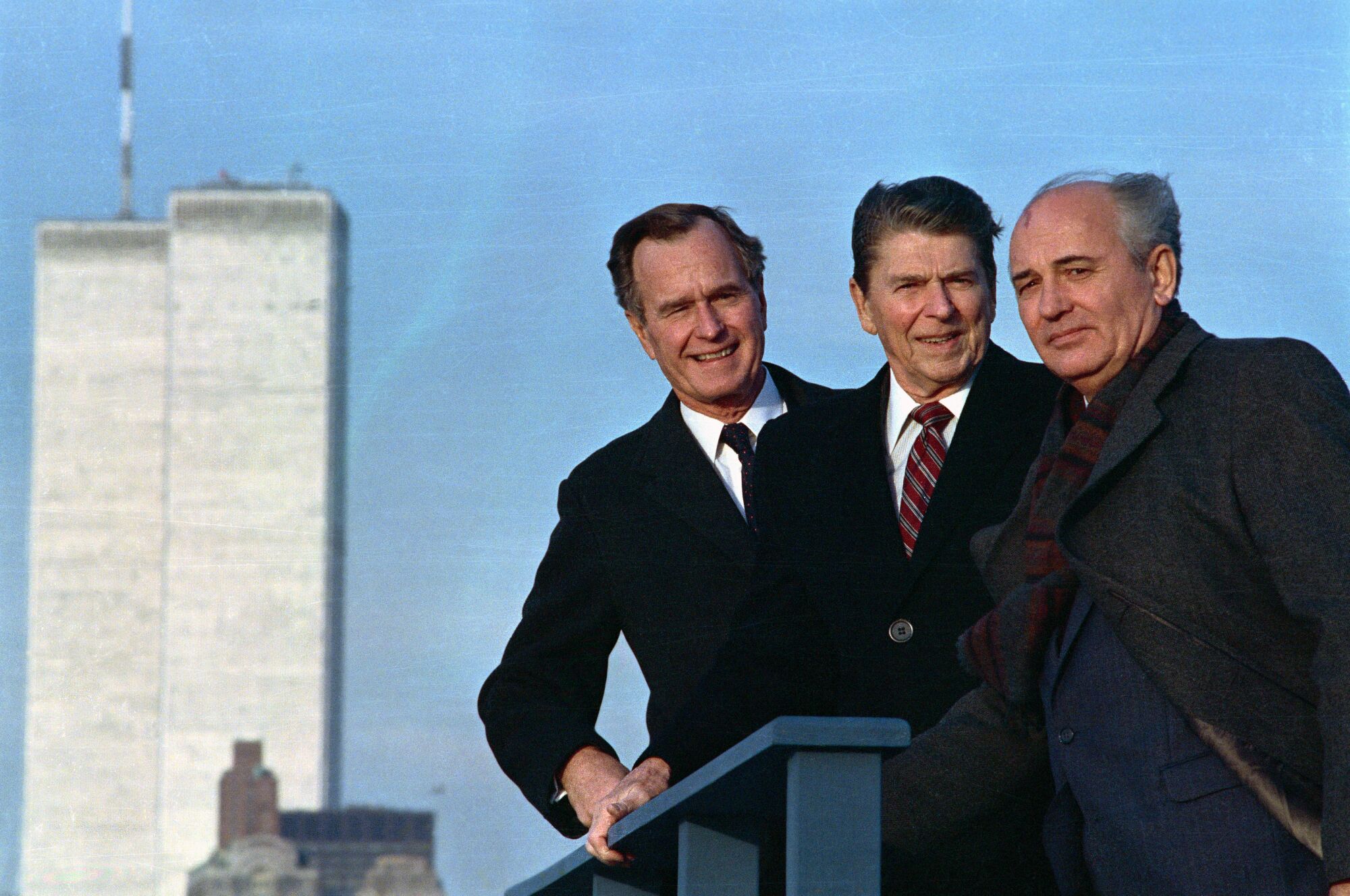Three men in dark suits stand near a blue railing, with two tall buildings as a backdrop 