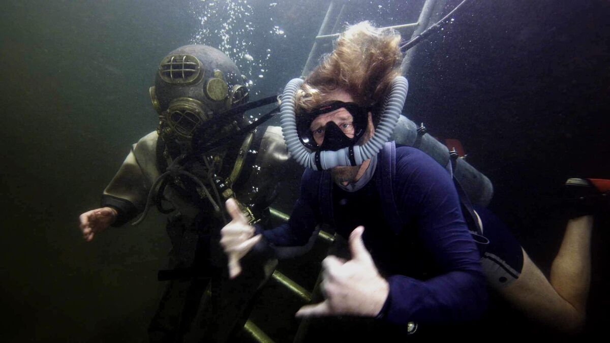 Two scuba divers underwater, one wearing an antique metal helmet and one making the "hang loose" shaka sign with both hands