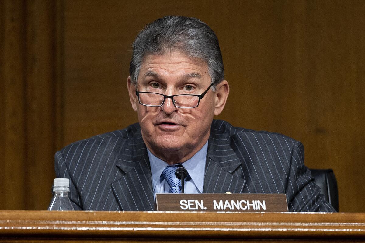 A man in a suit and glasses is seated behind a sign that reads Sen. Manchin 