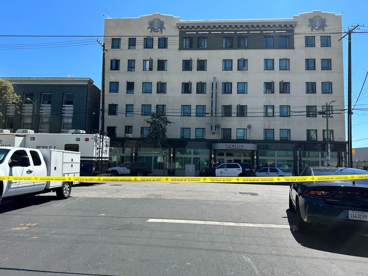 Police tape is seen in front of a parking lot. An older multi-story residential building is in the background.