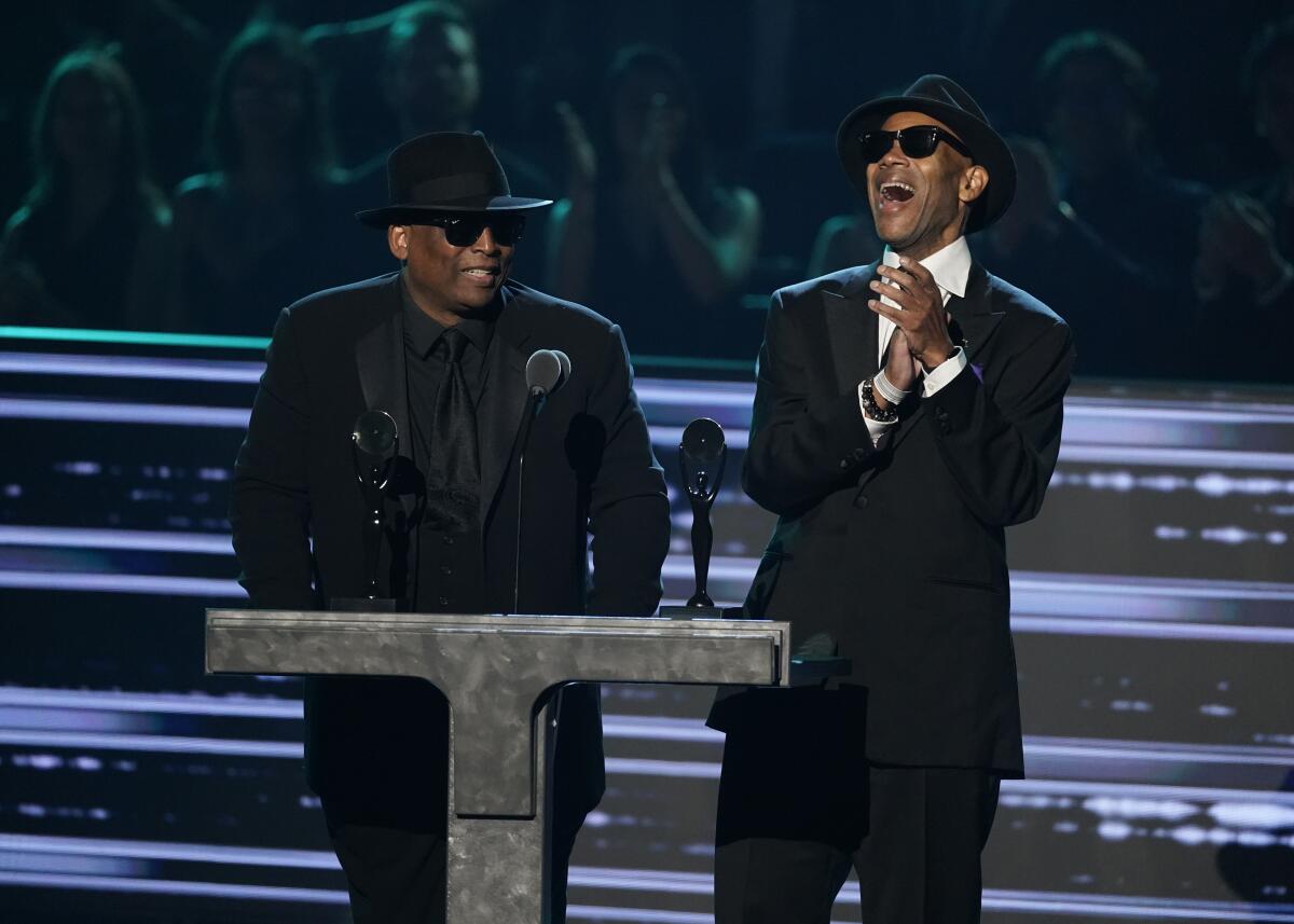 Two men in sunglasses and black hats speak from a podium