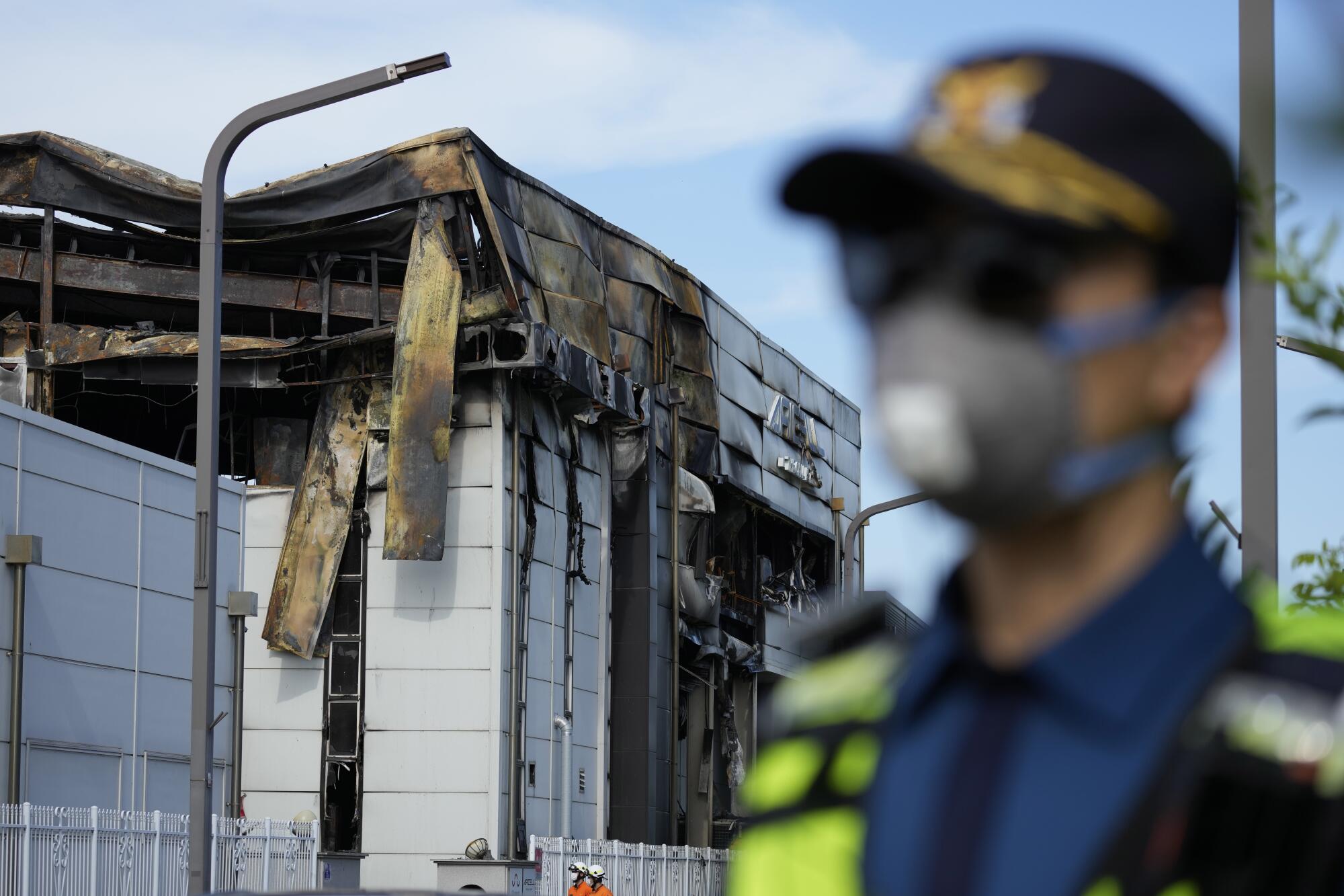 A police officer, out of focus in the front of the frame, stands near a scorched building.