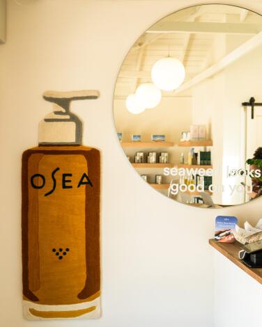 A round mirror reflecting a skincare studio on a wall next to a painted pump bottle labeled OSEA