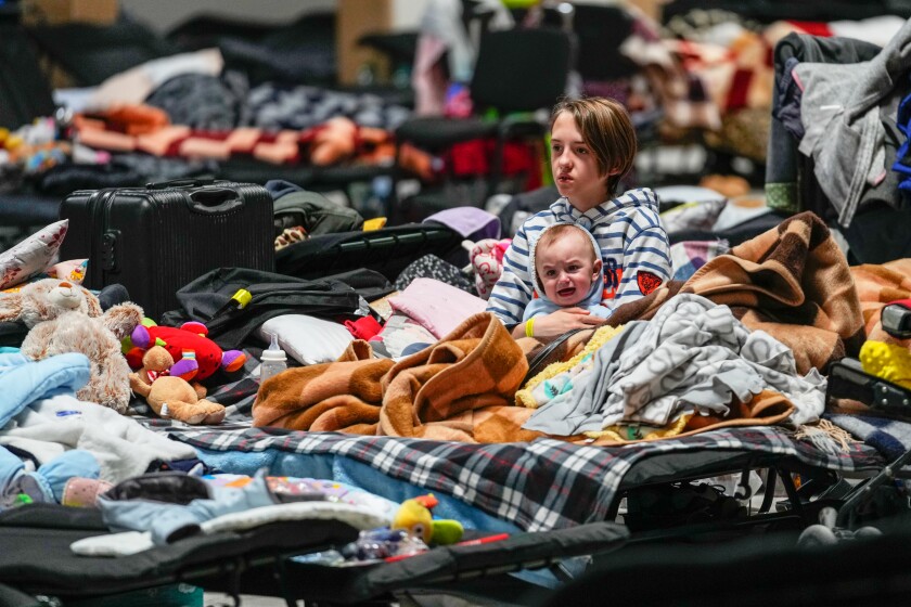An infant and an older child sit surrounded by blankets, toys and luggage