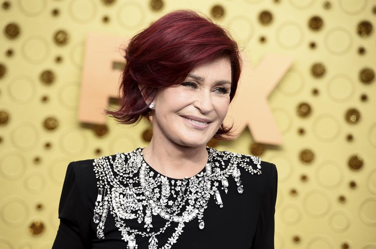 A woman with short red hair smiling in a bejeweled black dress against a gold background