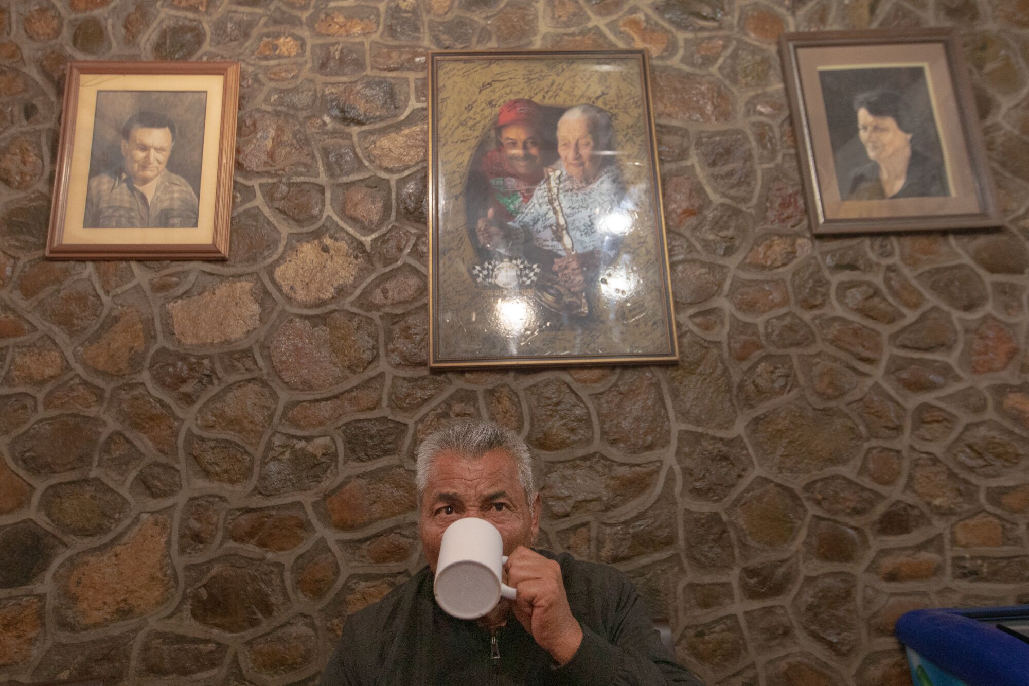 A man sipping coffee while sitting against a stone wall with three vintage-looking framed portraits.