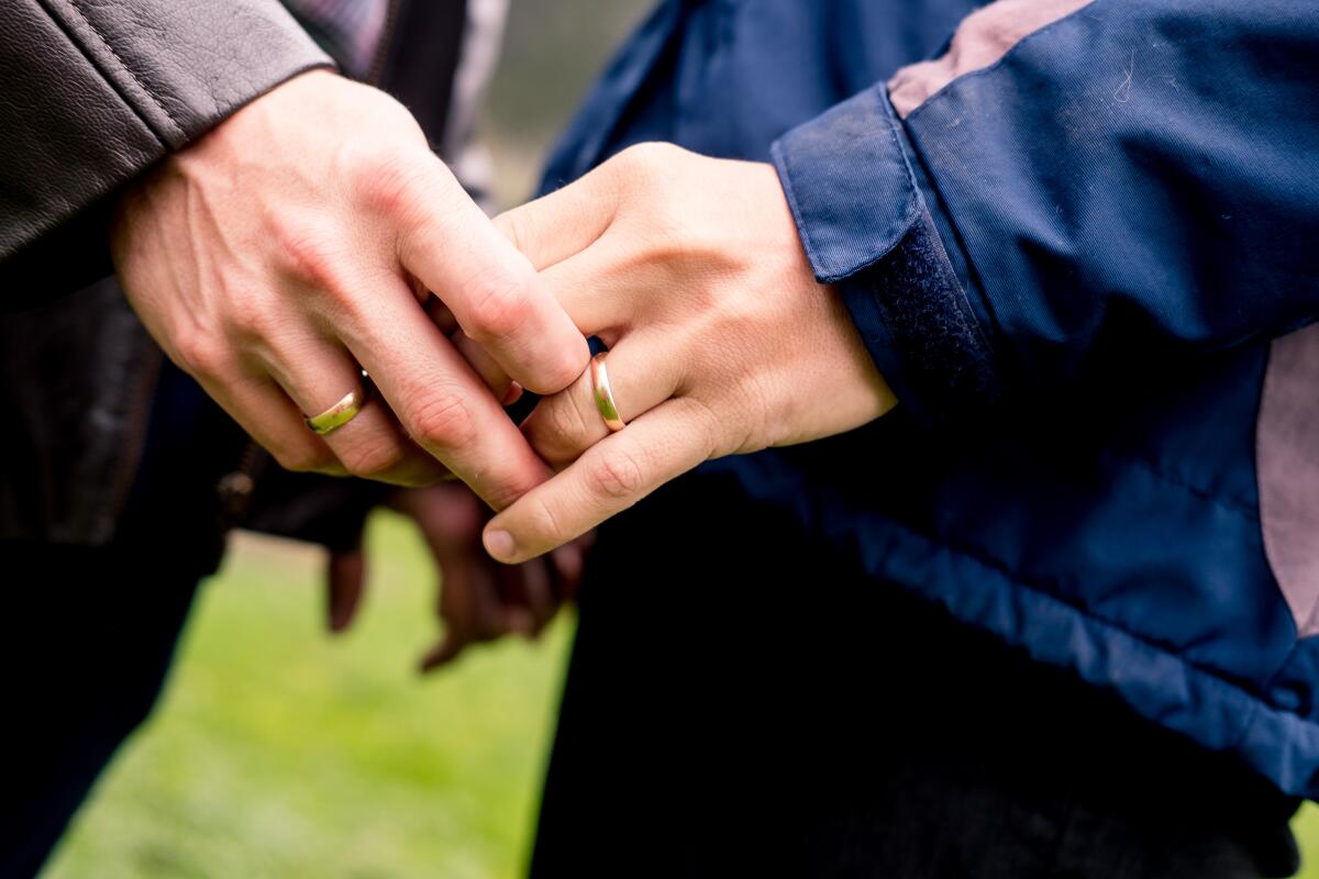 A close-up of a man and woman's entwined hands; each wears a gold wedding band.