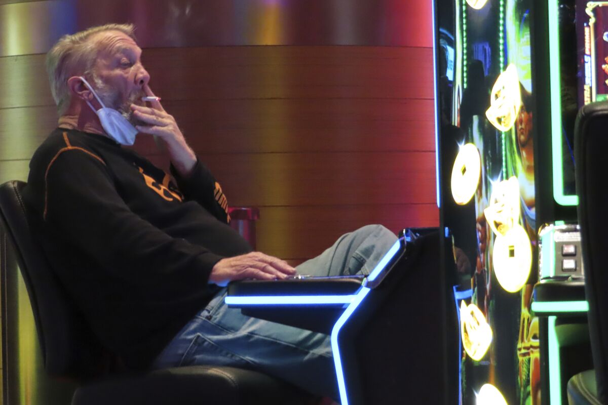 A man seated at a slot machine lowers his mask to smoke a cigarette.