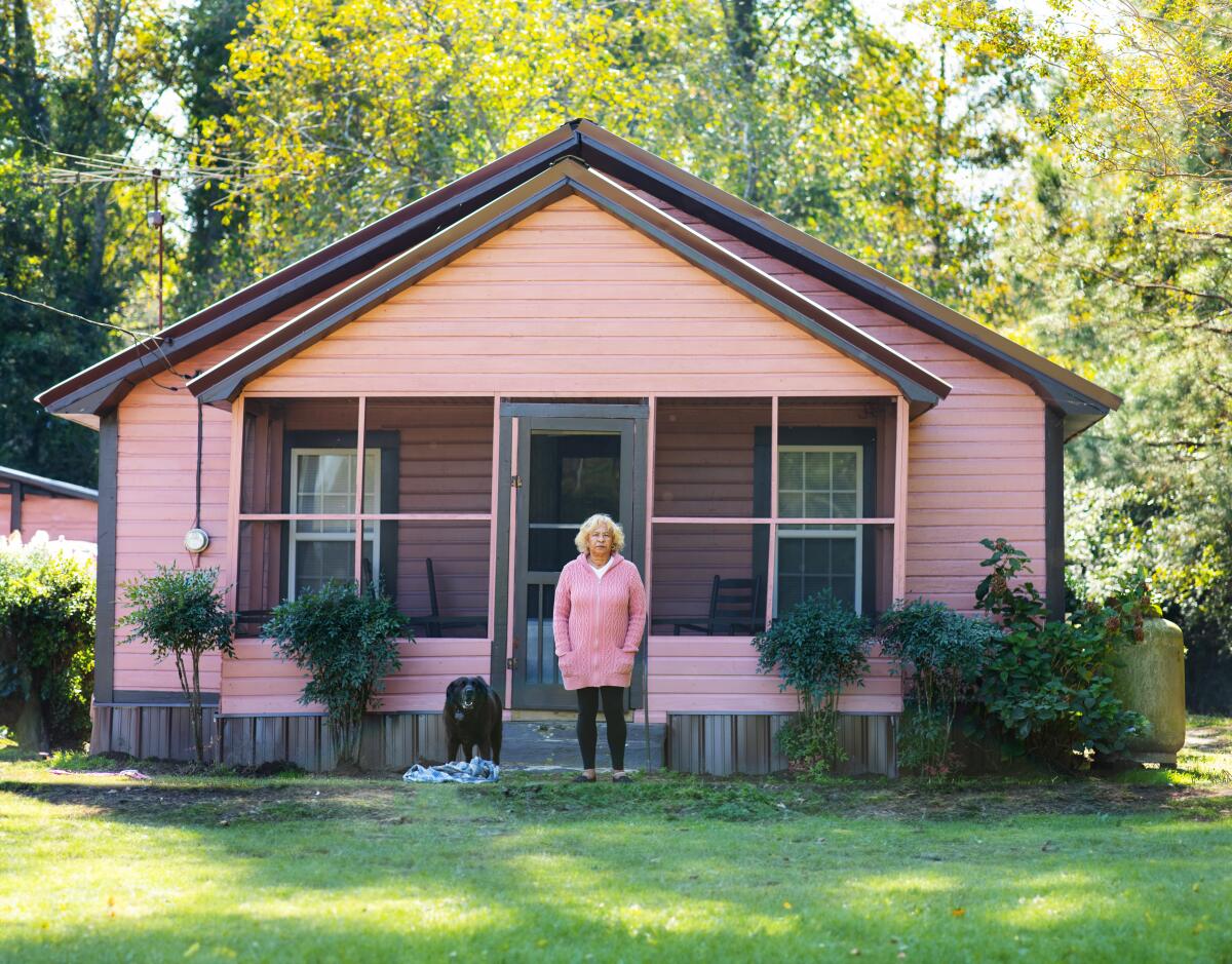 A woman in a pink top stands in front of her pink house with her dog.