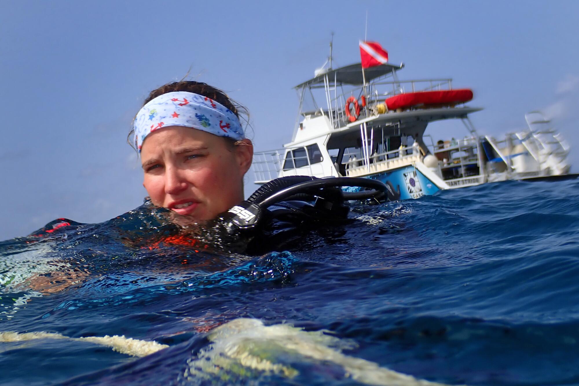 A woman surfaces from an ocean dive as a boat floats behind her. 