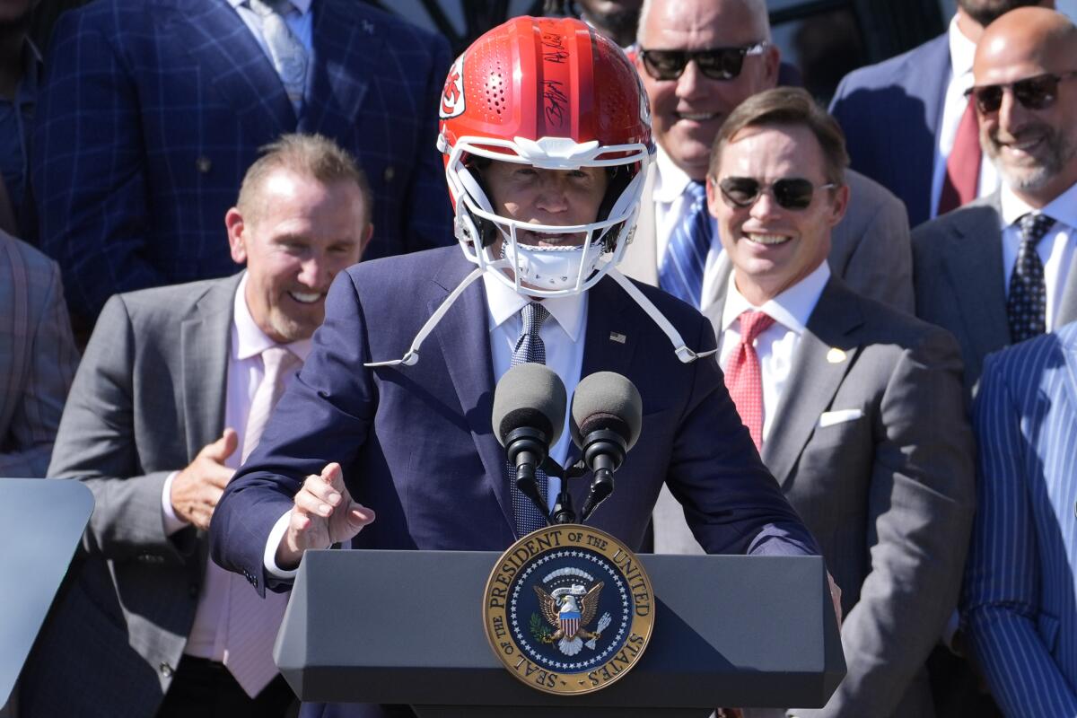 President Biden wears a Chiefs helmet at a dais, with the team smiling behind him.