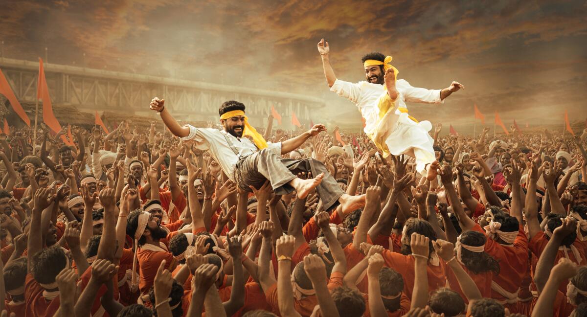 Looking joyous with arms in the air, Rama (Ram Charan) and Bheem (N. T. Rama Rao Jr.) are hoisted by a huge crowd 