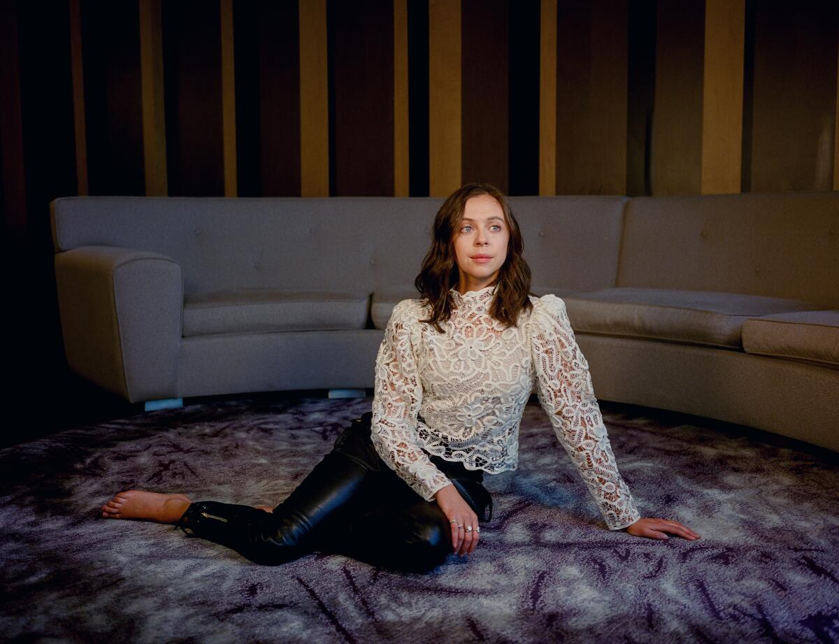 Bel Powley in a lace top and black leggings is sitting on a carpeted floor with her legs to the side.
