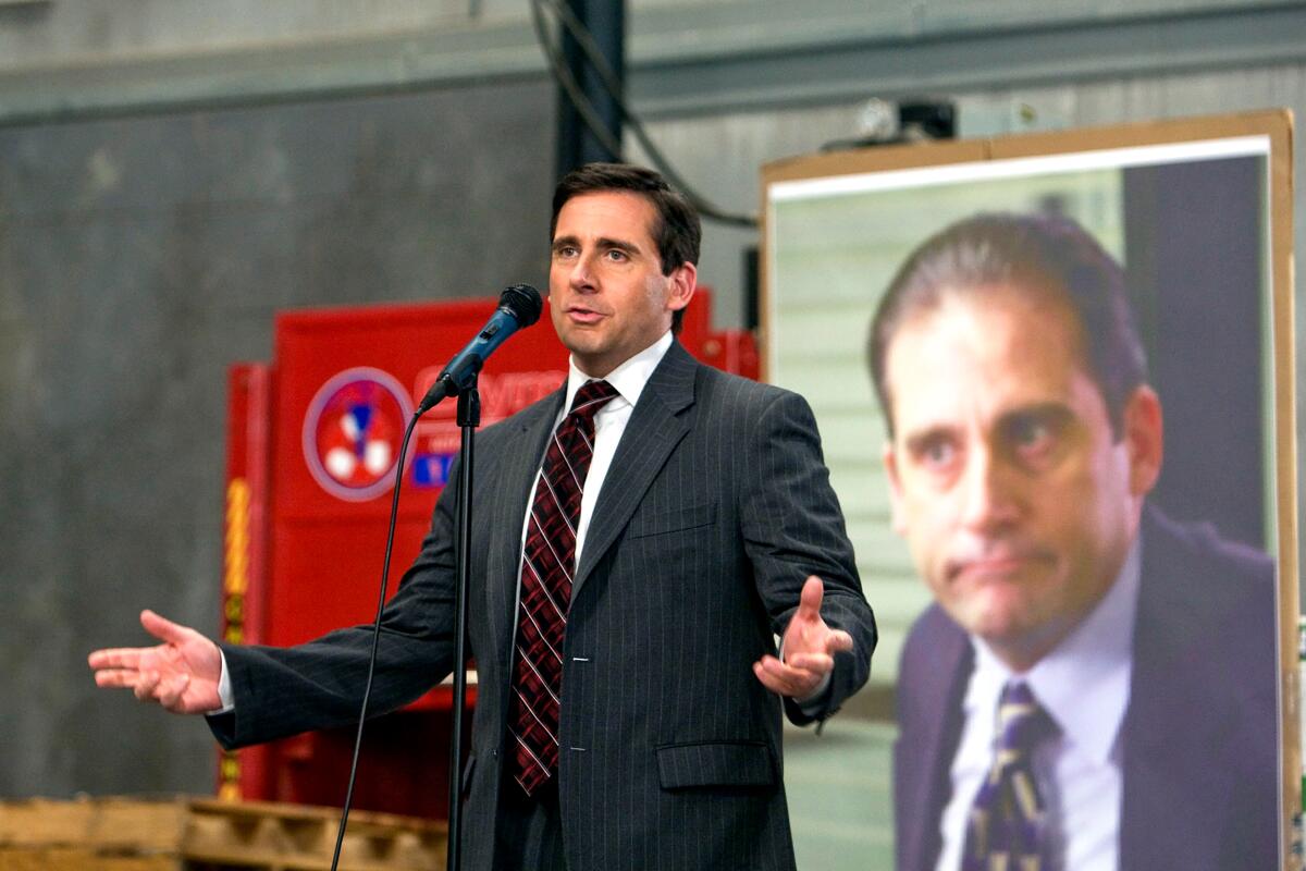 Steve Carell as Michael Scott, standing in front of a blown-up photo of himself, speaks into a mic in "The Office."