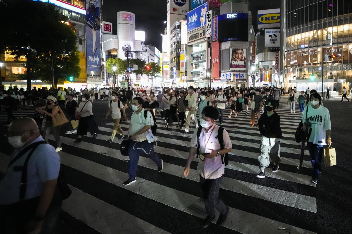 People wearing masks walk on a broad crosswalk with city lights and buildings behind them.