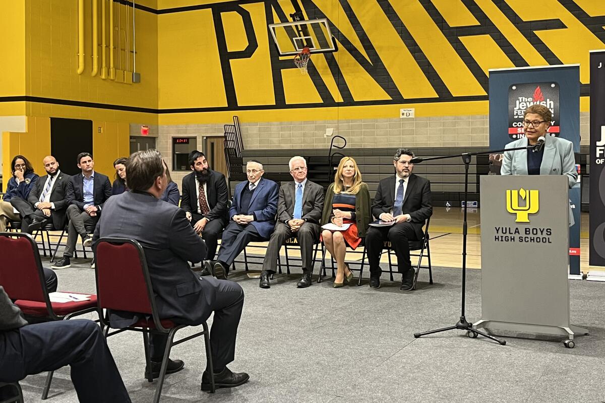 Karen Bass speaks at a lectern that reads "Yula Boys High School" in a yellow-painted gymnasium, while people sit on chairs.
