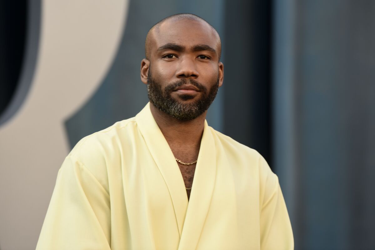 A bald man with a black beard posing in a loose yellow suit.