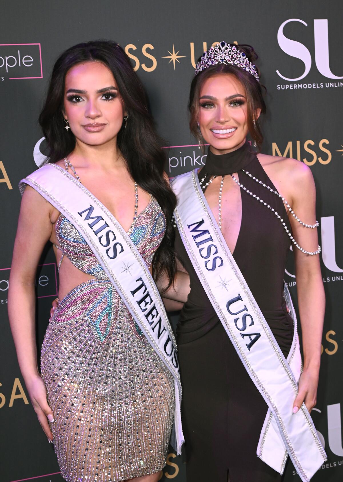 One pageant winner posing in a sash and short bejeweled dress with another winner clad in a sash, tiara, and black dress.
