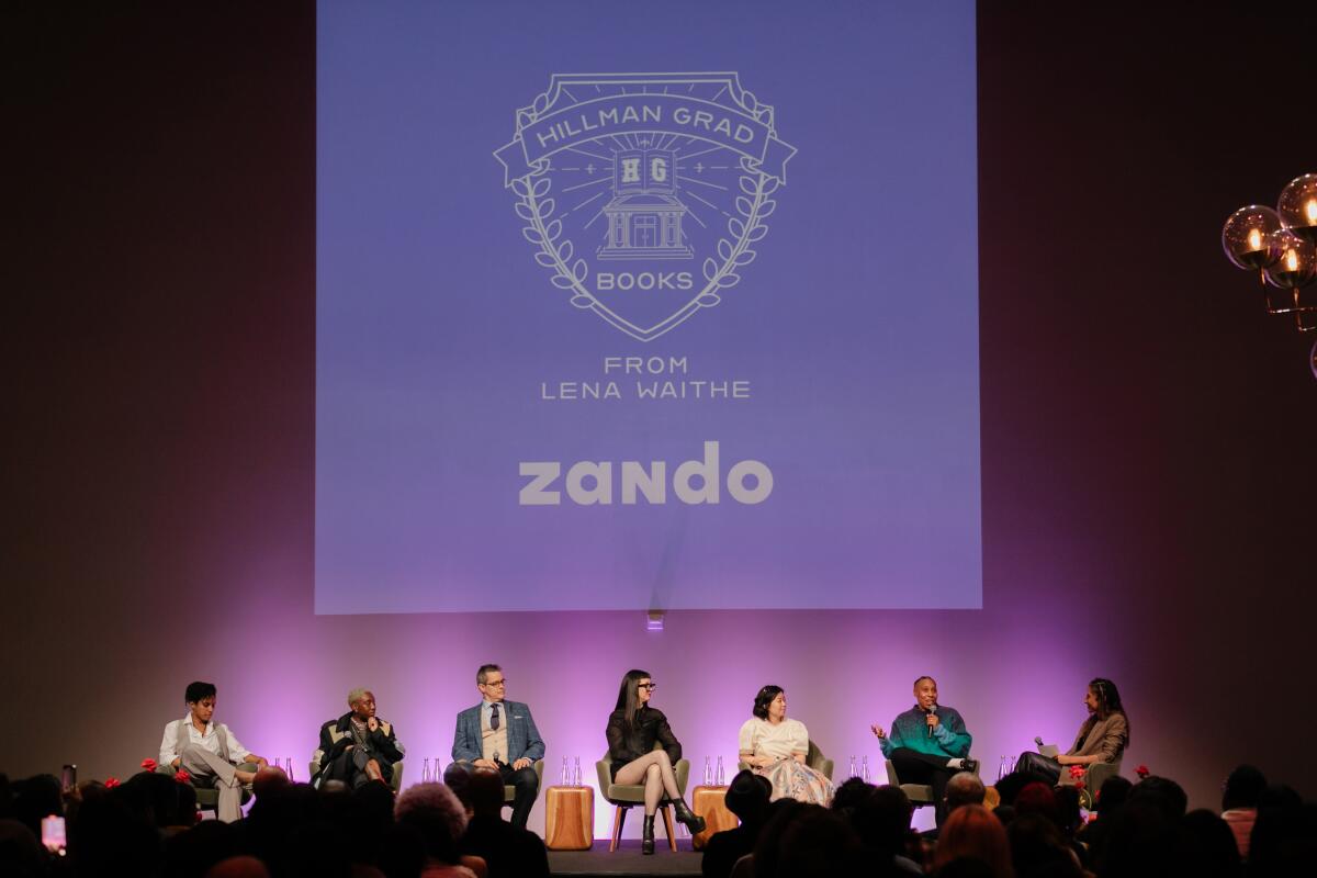 Seven men and women sit onstage in front of a purple projection with the words Hillman Grad from Lena Waithe and Zando.