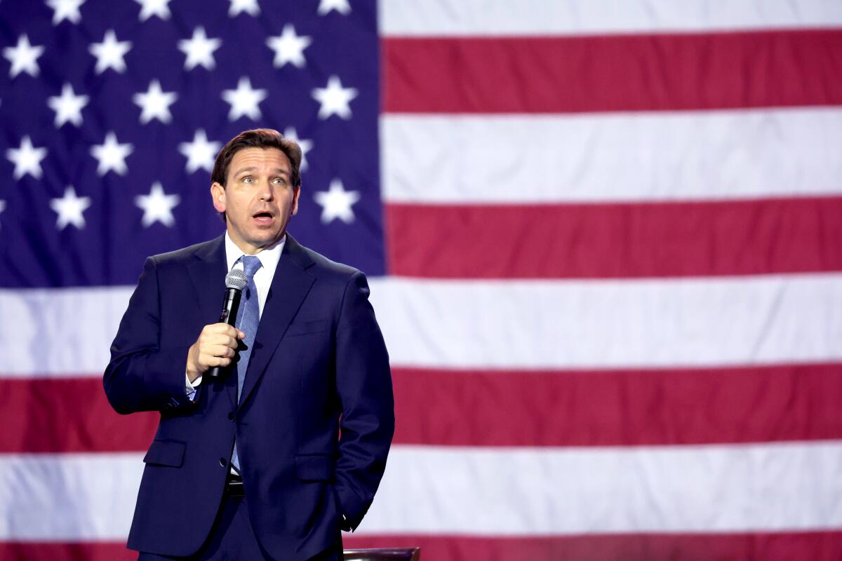 Ron DeSantis speaks into a microphone. A large American flag fills the backdrop.