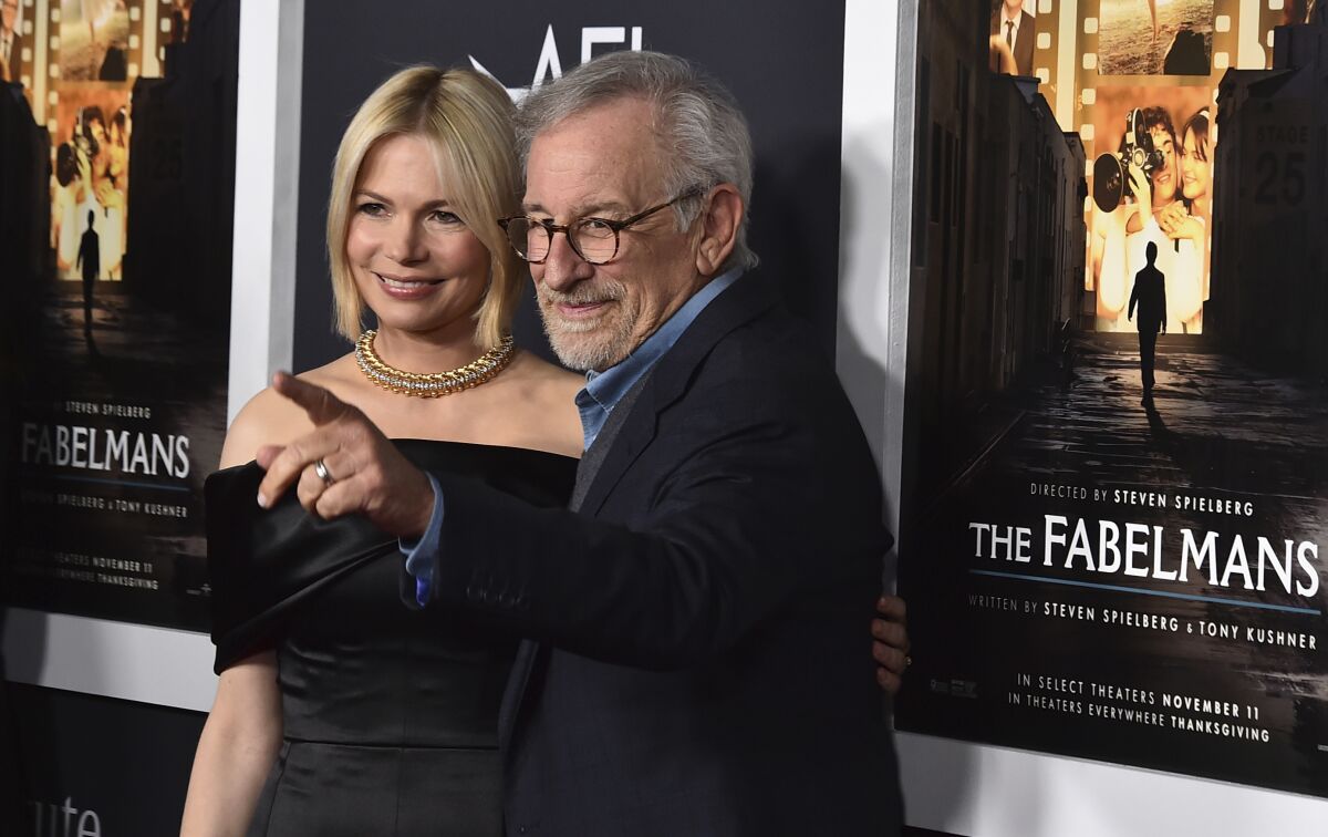 A blond woman in a black dress and a man with gray hair and glasses who is pointing in a black suit.