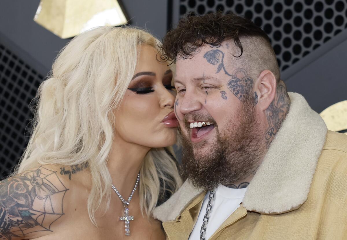 A woman with long blond hair and tattoos smooches the cheek of a man with facial tattoos and wearing a shearling jacket