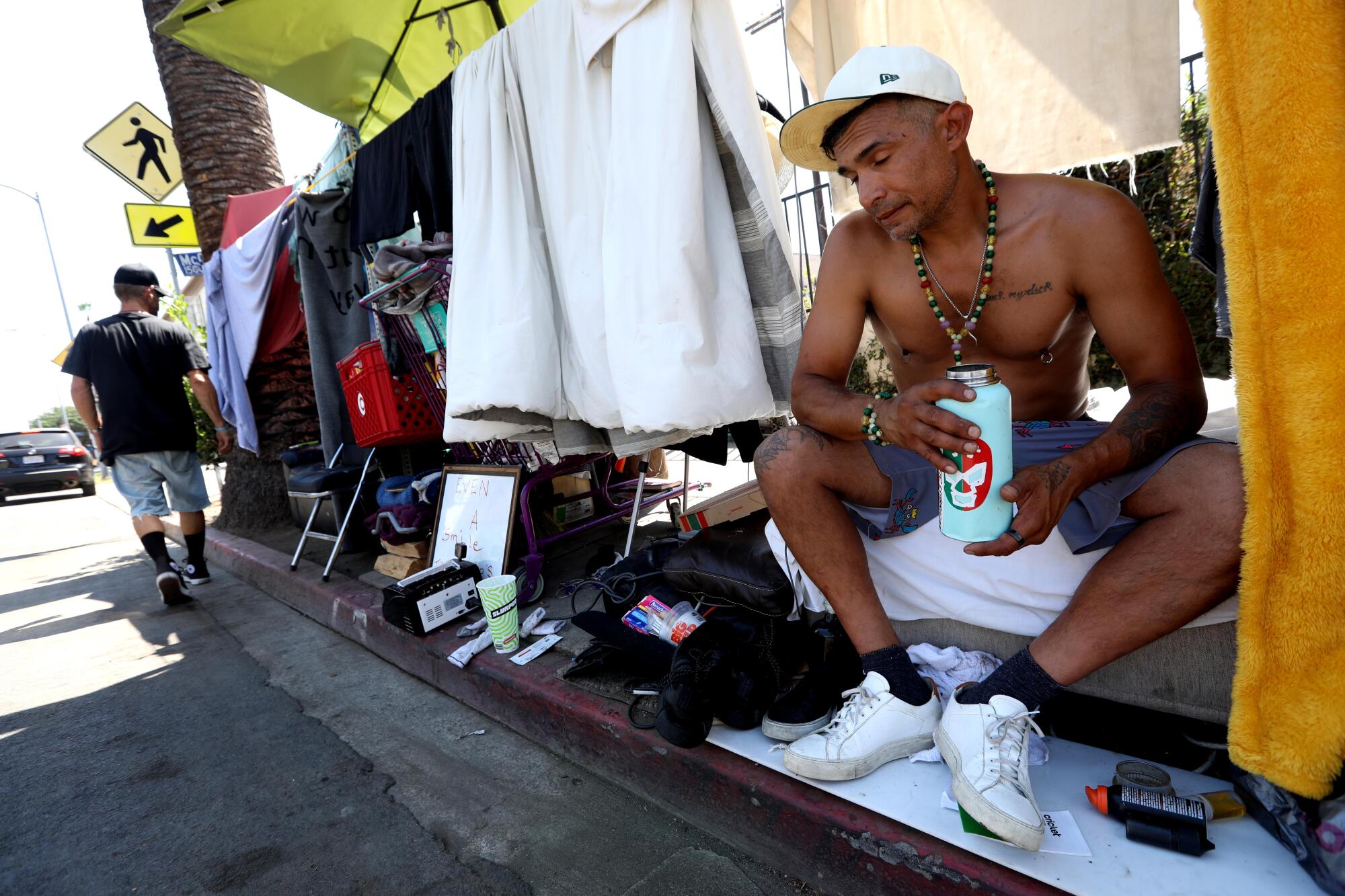 Shirtless man holds a drink while sitting in homeless encampment along the street. 