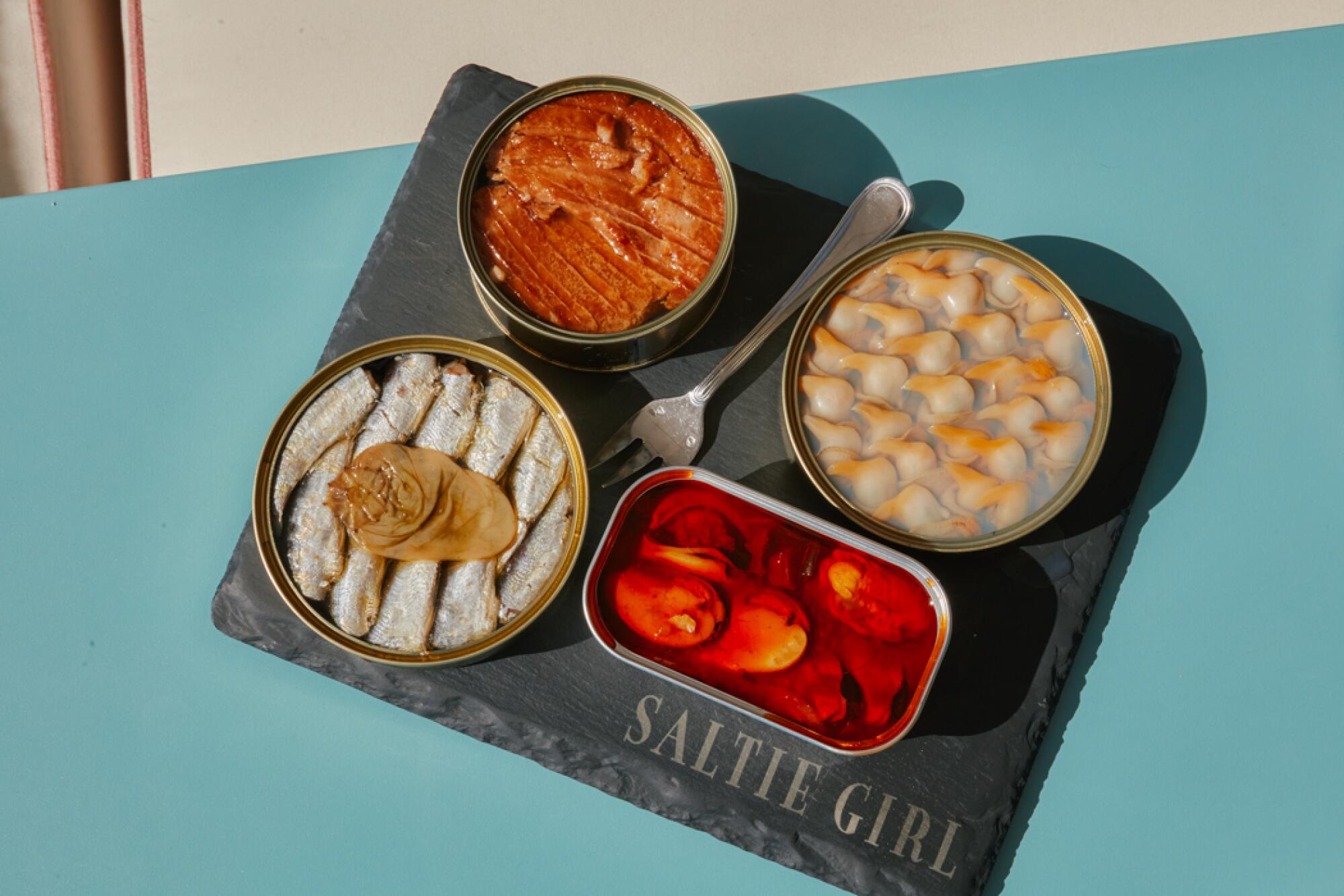 A view from above of four open tins of seafood with a fork on a slate board printed with the words Saltie Girl
