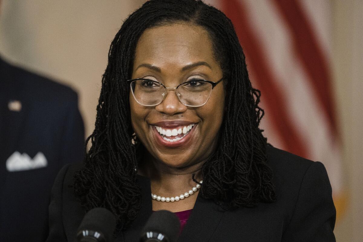 A Black woman wearing glasses and a dark jacket smiles as she speaks before microphones