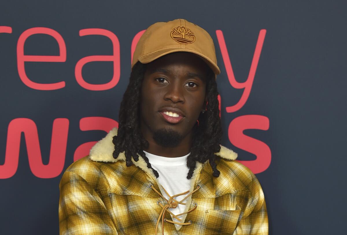 A young Black man with braids in a brown canvas hat and a yellow plaid jacket posing at a red carpet