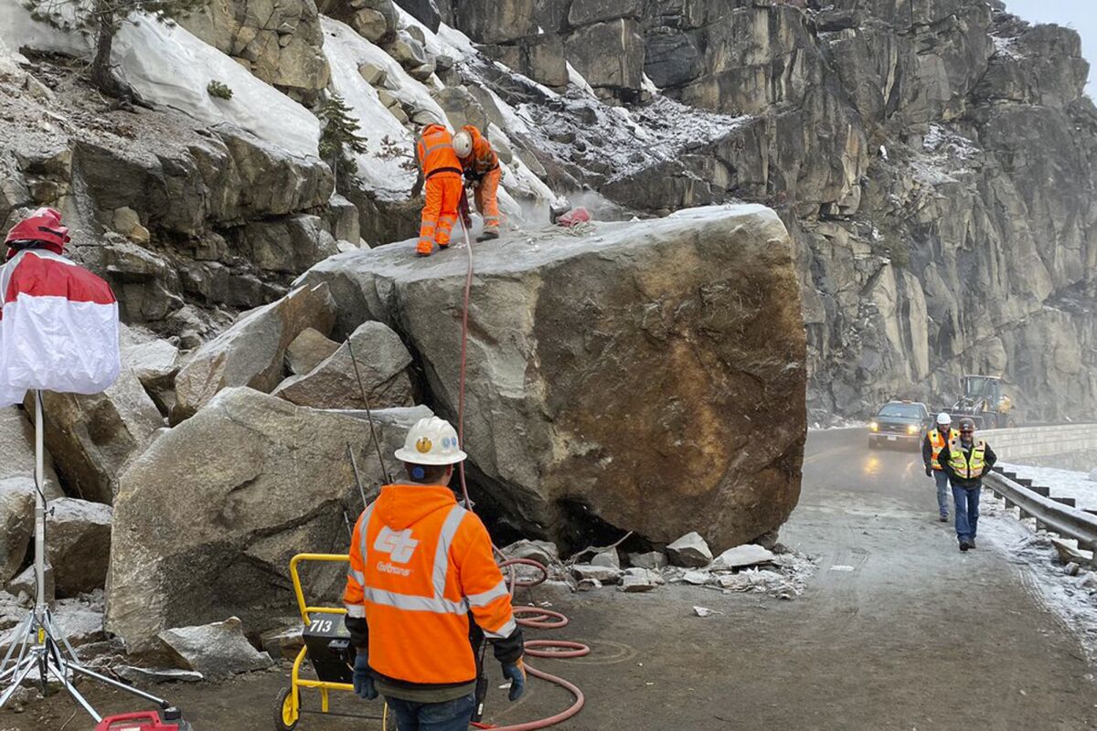In this photo provided by Caltrans, a crew drills holes to place explosives into a large boulder that fell onto U.S. Highway 50 near South Lake Tahoe, Calif., Friday, March 4, 2022. The return of winter weather prompted forecasters to advise people planning mountain travel to be prepared for snowy roads Friday and Saturday. Travel through the Sierra was disrupted by the massive boulder that fell onto the highway at Echo Summit on Thursday. (Caltrans via AP)