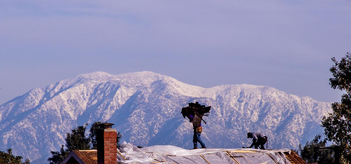 A person carrying a load of shingles of their head walks across a roof. Mountains are in the background.