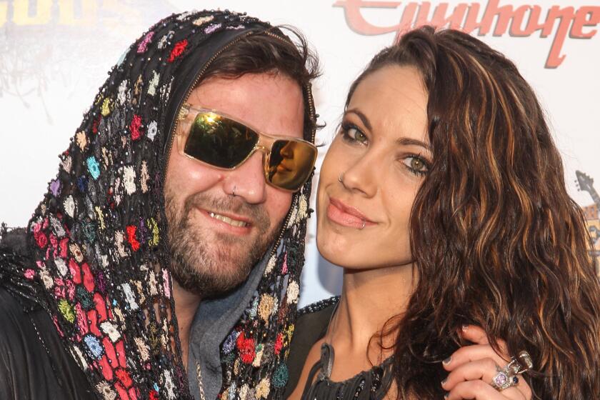 A man in sunglasses and a brightly colored headscarf poses at an awards show with an attractive woman with long brown hair 