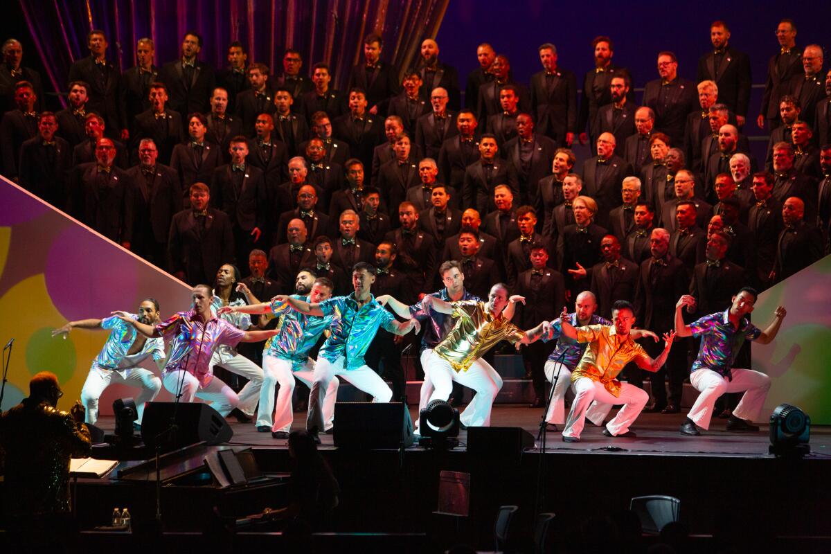 A large chorus of people onstage with some performers wearing colorful shirts.