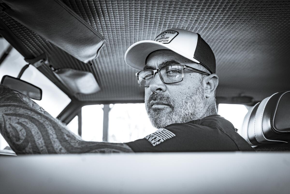 A man in a trucker cap and tattoos sits in an old car looking out the side window