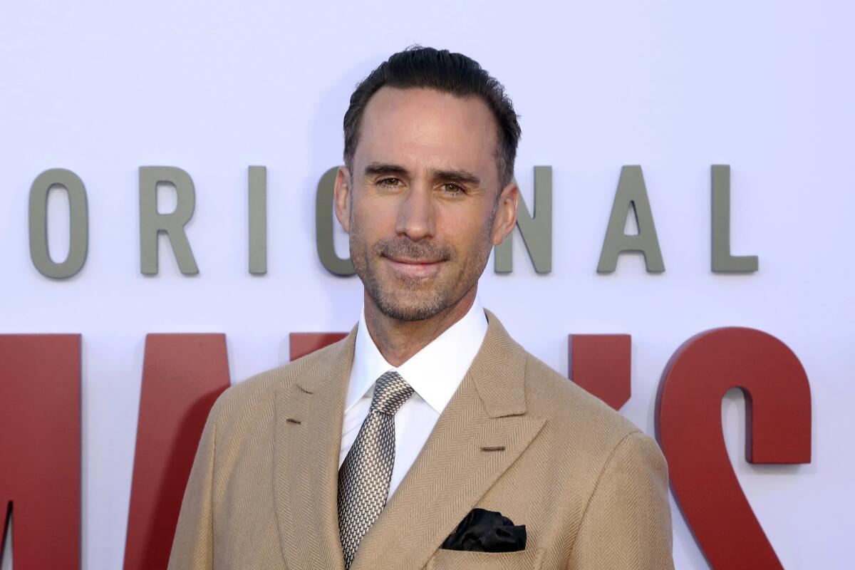 Joseph Fiennes smiling in a tan suit, white suit shirt and silver patterned tie