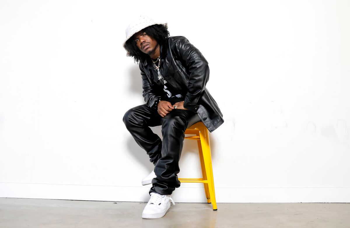 Rapper in white bucket hat dressed in black sitting on a stool against a white wall