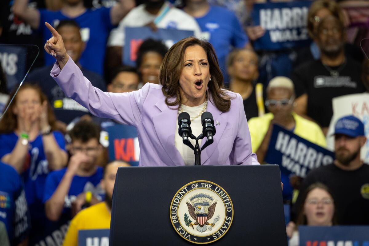 Kamala Harris points up and speaks from behind the vice presidential seal as people in the background raise campaign signs