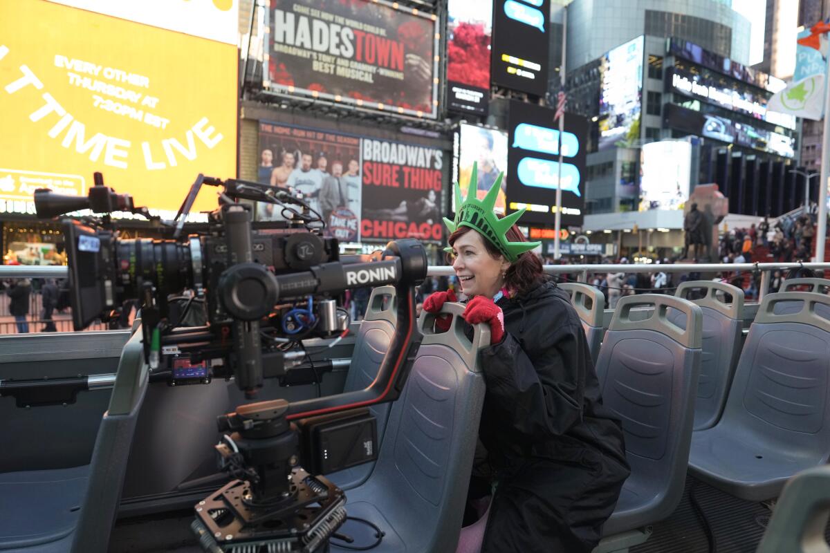 A woman in a Statue of Liberty crown rides in an open-top bus through Times Square, a TV camera filming her