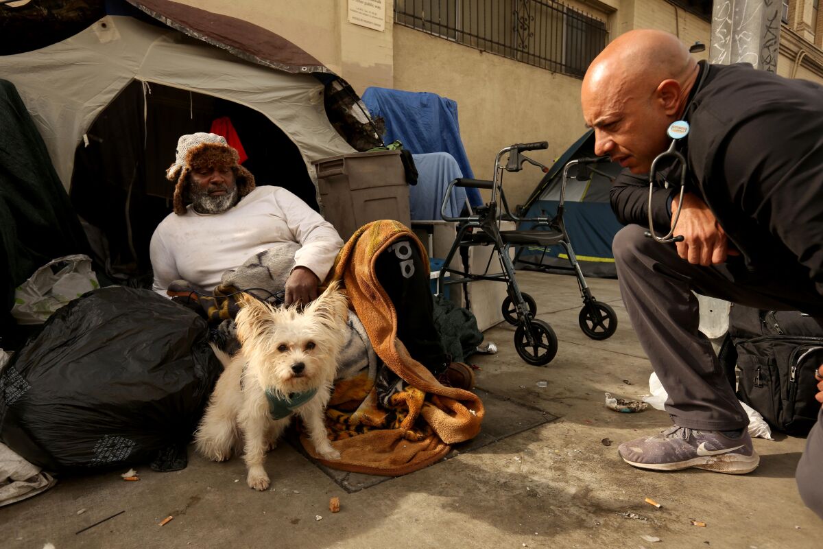 A man with a stethoscope kneels next to a man and small dog on a sidewalk in front of a tent.