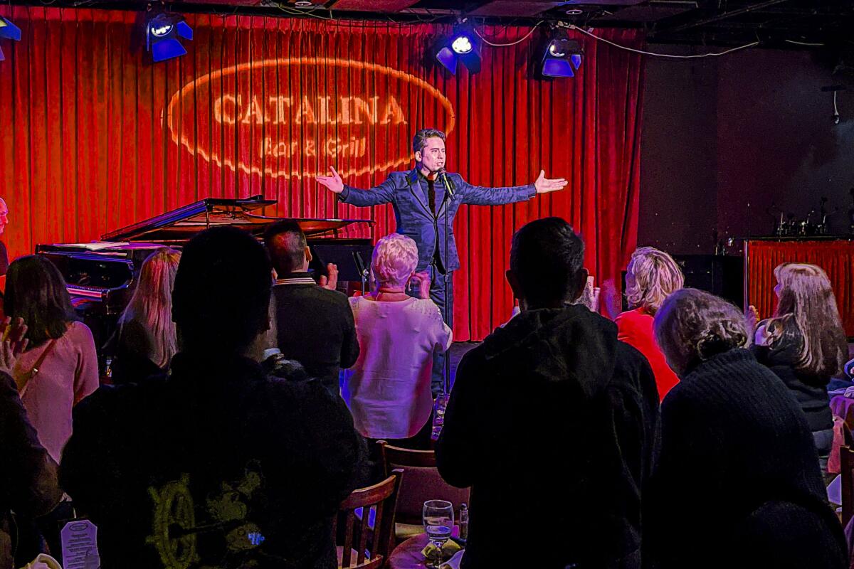 A man onstage in a small club next to a piano, the crowd standing to applaud him