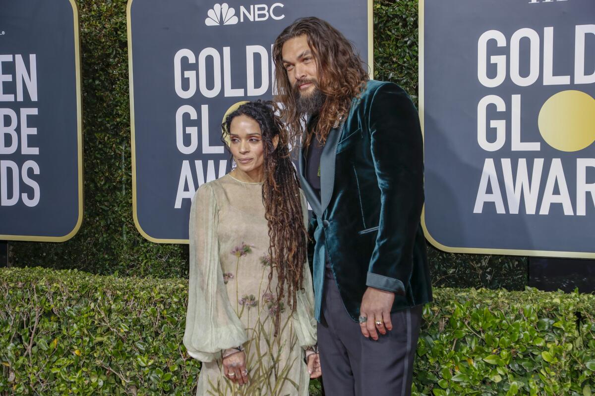 Lisa Bonet wearing a sheer green dress with floral pattern poses next to Jason Momoa, in a velvet green suit jacket.