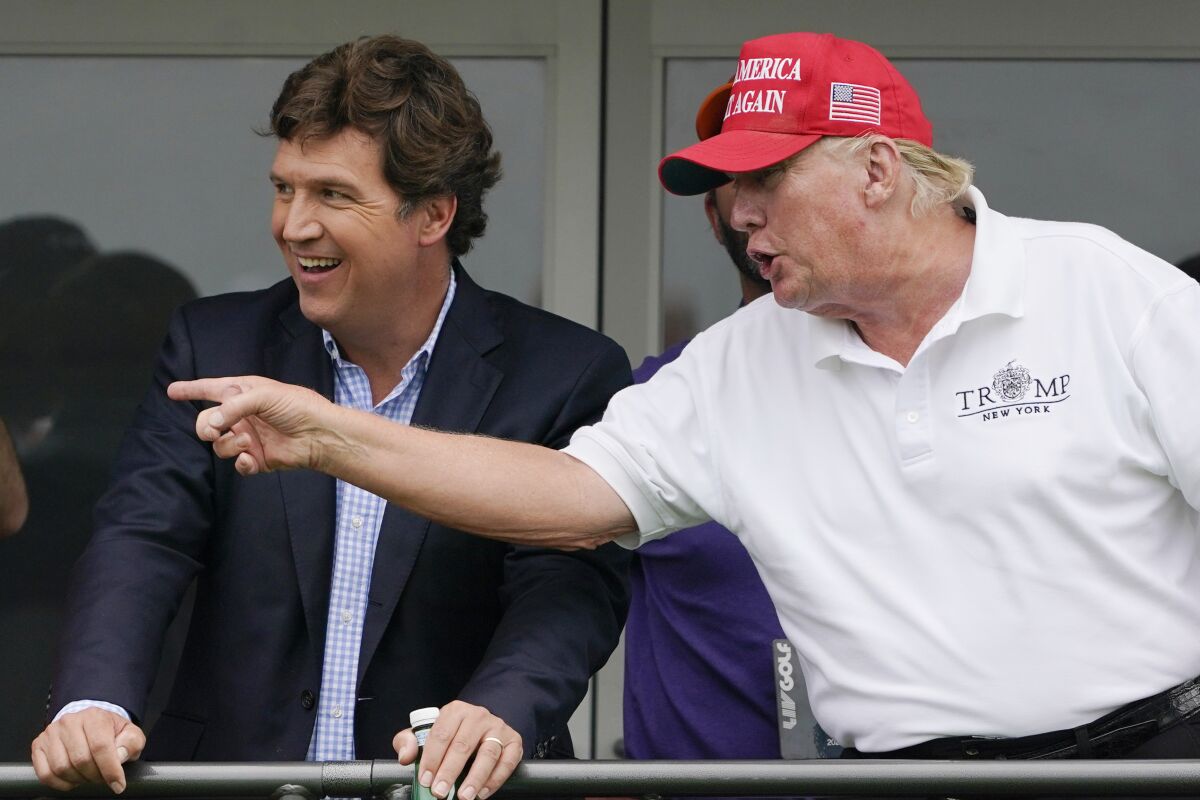 Donald Trump, in a "Make America Great Again" hat, gestures and Tucker Carlson looks.