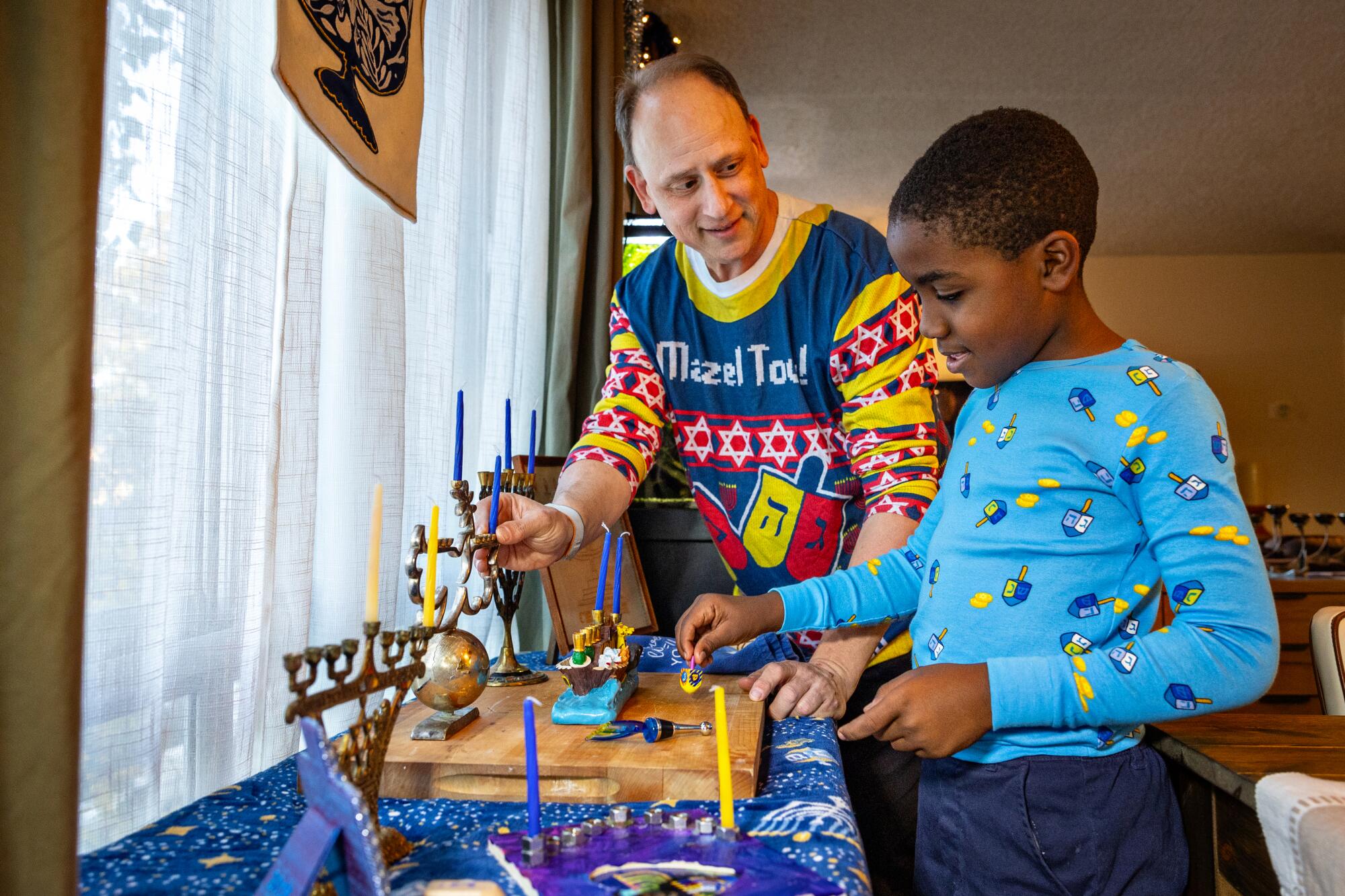 A man and a boy stand near menorahs at a window.