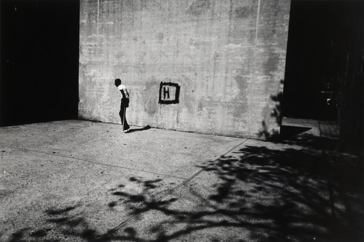 A black-and-white photo of a young man near a gray wall that features the painted outline of a rectangle with an H inside it.