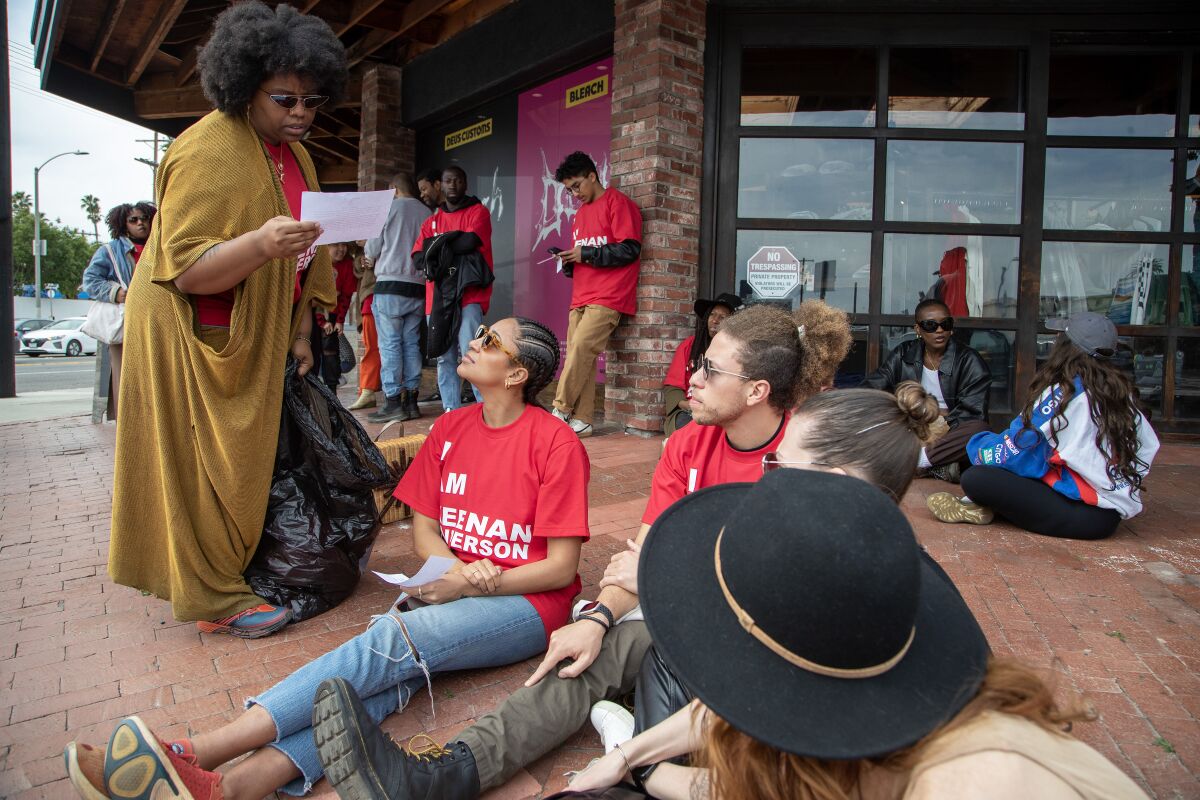 Patrisse Cullors stands lookimg at a document while other protesters sit on a sidewalk.