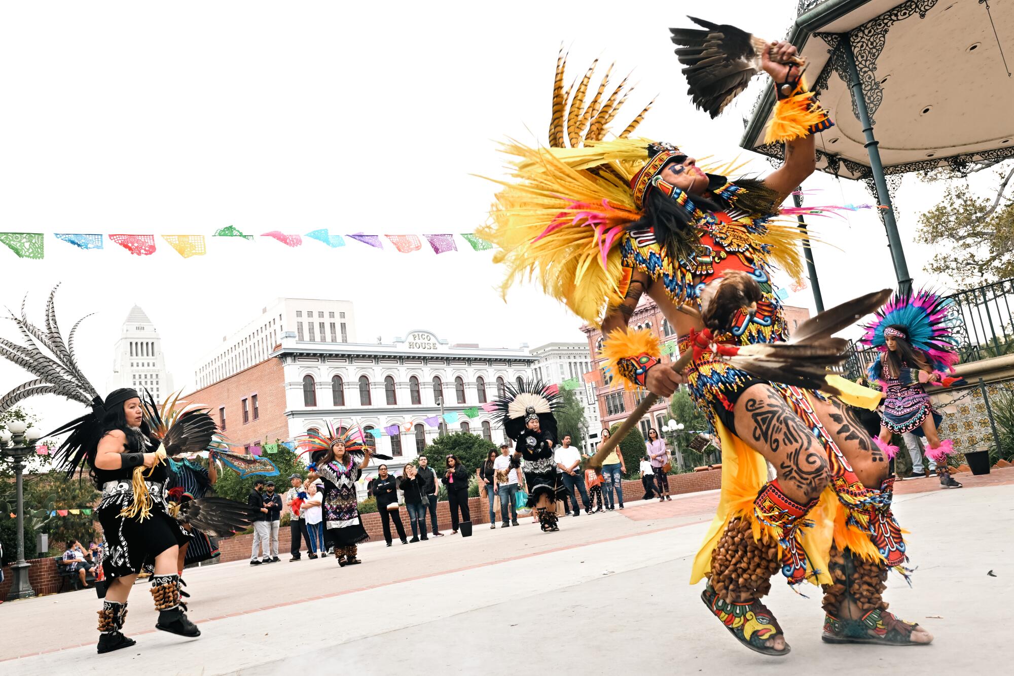 Dancers wearing feathered outfits perform in a courtyard while a group of people looks on.